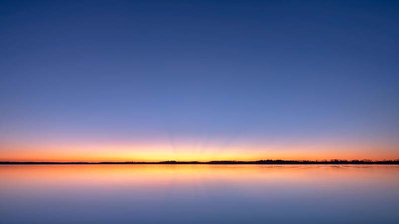 A calm lake reflects a deep purple sky with vibrant red-orange colors on the horizon at sunset