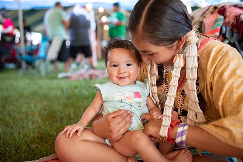 An Oneida woman sits on the ground and holds her smiling infant daughter