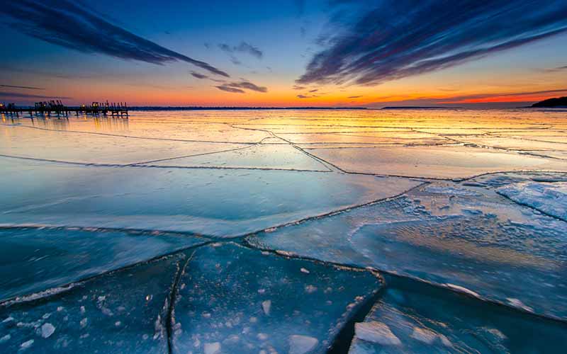 An ice-covered lake with numerous cracks reflects the brilliant colors of a winter sunset