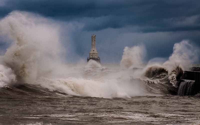 Winds whip up waves that crash against the lakeshore as a lighthouse stands against a blue-gray sky