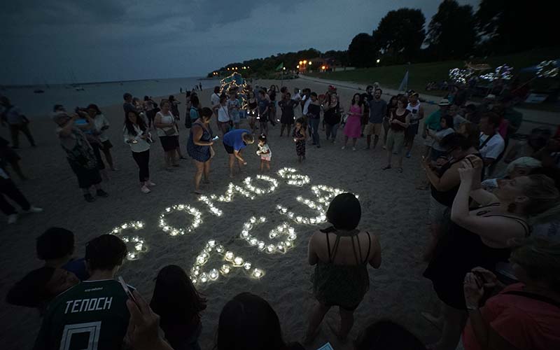 Community members gather at dusk on a beach around candles arranged to spell 