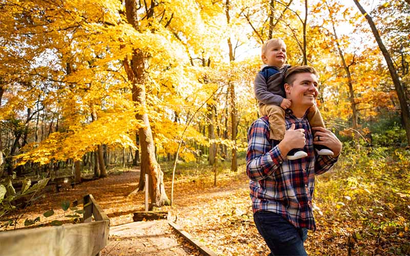 Bright fall colors surround a young father and his toddler son as they walk through a forest