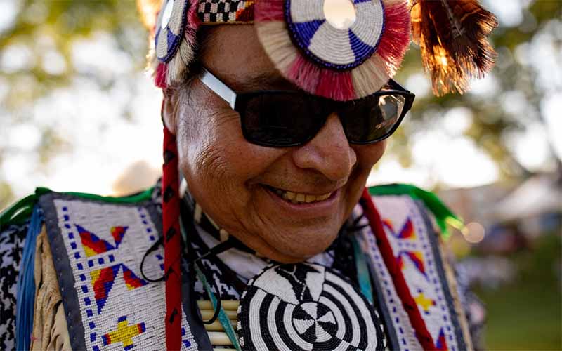 Closeup portrait of a member of the Oneida Nation, dressed in traditional clothing, smiling and wearing sunglasses