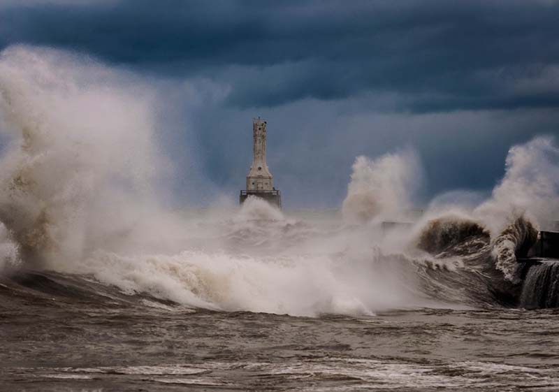 Winds whip up waves that crash against the lakeshore as a lighthouse stands against a blue-gray sky