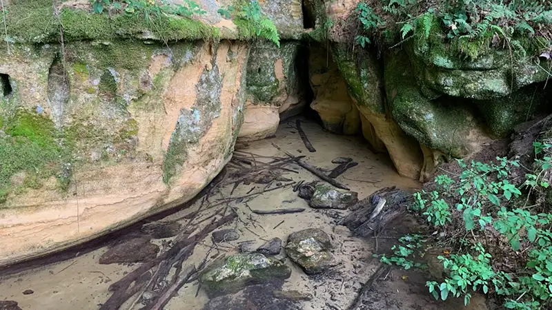 A shallow, sandy pool of water sits at the base of moss-covered sandstone rock formations and a small cave opening.
