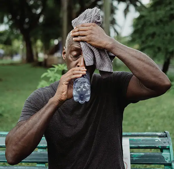 A man sits on a park bench, drinking water and holding a towel to his head to cool down during extreme heat.